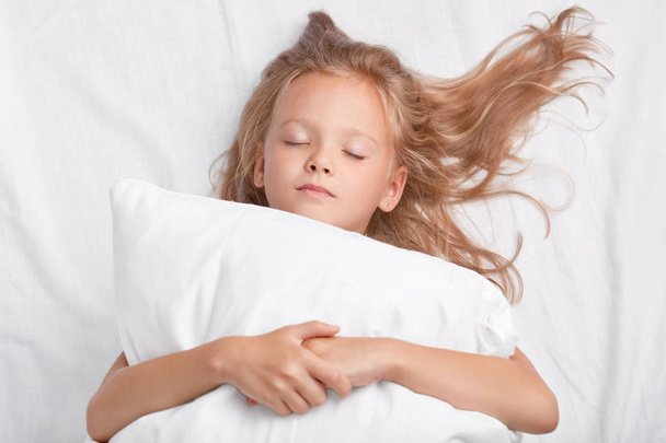 Little attractive daughter with closed eyes, embraces soft white pillow during sleep, enjoys domestic cozy atmosphere, lies in white bed, being photographed by mother. Bedding and rest concept - Fotografie, Obrázek