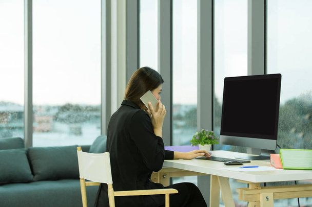 黒いスーツの若いアジア女性実業家は、スマート フォンを呼び出しているし、机の上にコンピューターがある窓が大きくて近代的なオフィスに座っています。. - 写真・画像