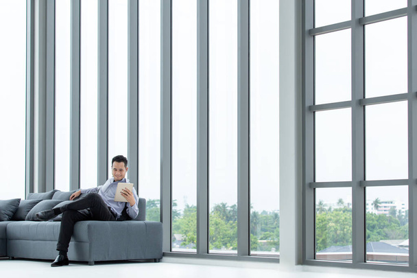 Молоді азіатські бізнесмен погляд на планшет, сидячи на дивані щасливою усмішкою. Великі вікна в сучасному офісі. - Фото, зображення