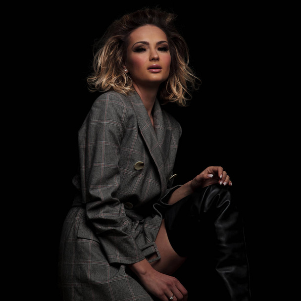 portrait of beautiful blonde woman in grey jacket with plaids resting hand on thigh while standing on black background with parted lips - Photo, Image