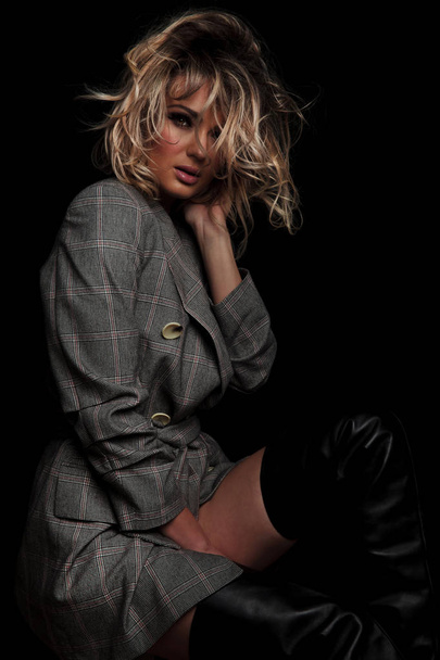 sensual blonde woman in grey plaid jacket sitting with hand between her thighs while fixing her hair on black background - Zdjęcie, obraz