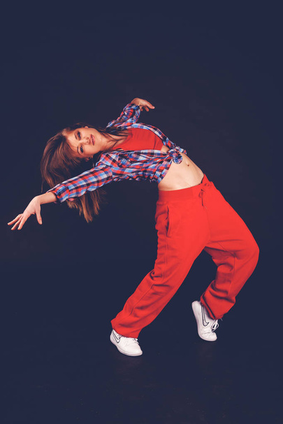 Young beautiful woman girl in red pants and plaid shirt dancing hip hop on a dark background - Foto, Bild