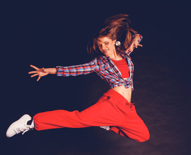Young beautiful woman girl in red pants and plaid shirt dancing hip hop in jumping against a dark background - Valokuva, kuva