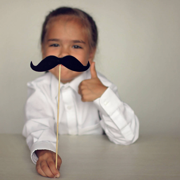 A cute little girl holding a fake felt moustache in a stick in front of her face, happy father day and men health concept - Photo, Image