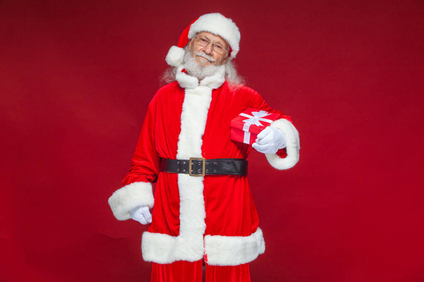Vánoce. Usmívající se Santa Claus v bílých rukavicích drží červené dárkové krabičce s mašlí. Ukázal na dárek. Izolované na červeném pozadí. - Fotografie, Obrázek
