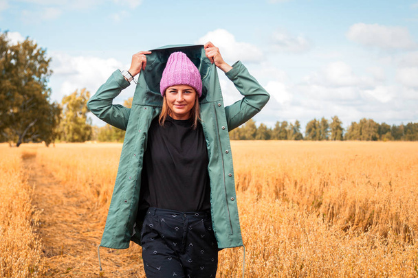 Jonge vrouw met groene vacht, hoed, jeans, wandelen, genieten van natuur en zonlicht in stro veld breien. Concept van de herfstvakantie bij dorp en levende stijl - Foto, afbeelding