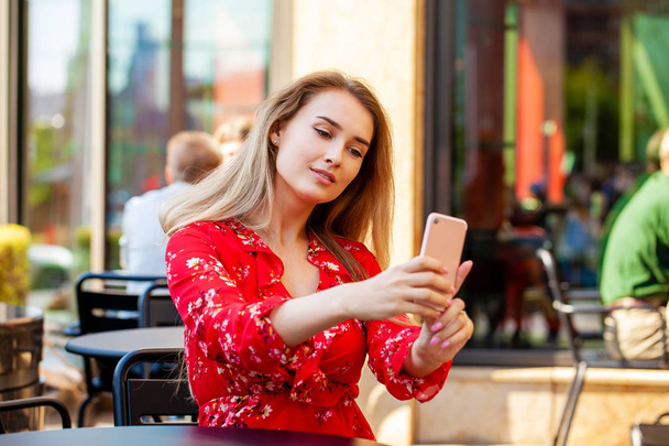 Young beautiful blonde woman in red dress taking pictures of herself on a cellphone sitting in a summer cafe on the street - Foto, afbeelding