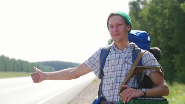 Egy fiatal férfi kockás ing, egy zöld kalap és egy hátizsákban állt az út, és honking autók - Felvétel, videó