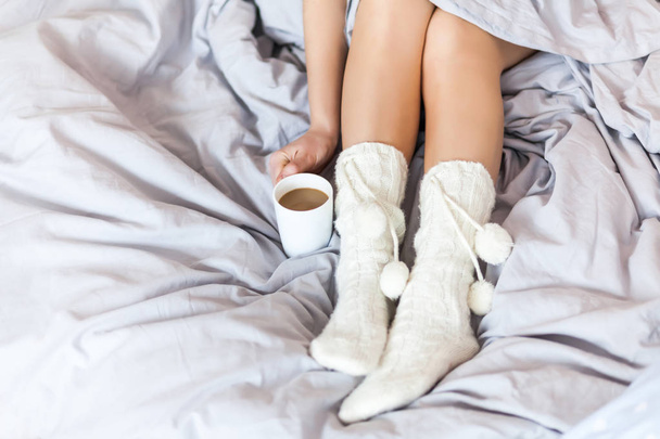 Woman resting keeping legs in warm socks on bed with morning coffee and reading book - Foto, Bild