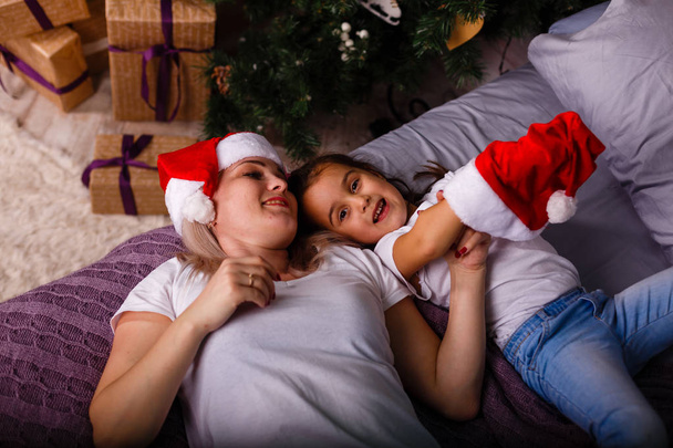 Chica feliz y madre sonriente pasar tiempo juntos cerca del árbol de Navidad
 - Foto, Imagen