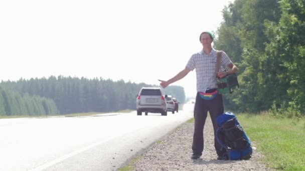 Um homem com uma camisa xadrez, um chapéu azul, com um acordeão no ombro ao pé da estrada e buzinando carros passando, deitado ao lado da mochila
. - Filmagem, Vídeo