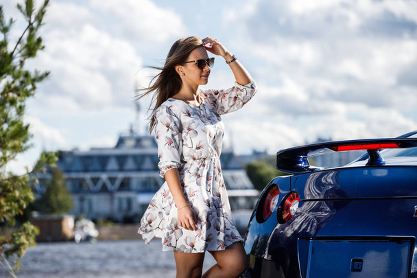 Outdoor shot of beautiful young woman in white dress in flowers. Female model standing close to modern sport car on street at daytime - Photo, image