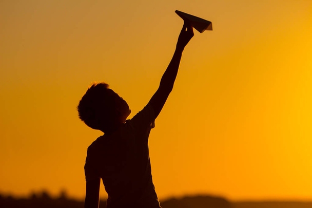 little boy holds a paper airplane in his hand at sunset. A child raised his hand up to sky and plays with origami in the evening in the street - Photo, Image