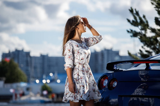 Außenaufnahme einer schönen jungen Frau in weißem Kleid und Blumen. Model steht tagsüber neben modernem Sportwagen auf der Straße - Foto, Bild