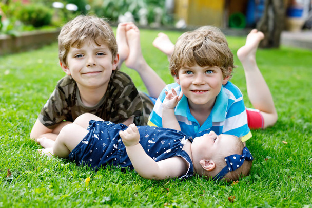 Two little happy kid boys with newborn baby girl, cute sister. Siblings on grass in summer or spring in garden. Kids bonding. Family of three children. - Photo, Image
