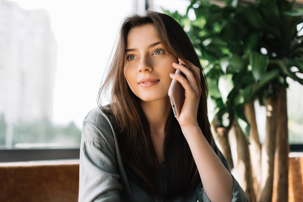 Closeup portrait of attractive caucasian business woman holding smartphone  sitting in cafe. Young beautiful female using mobile phone, working from home - Photo, image