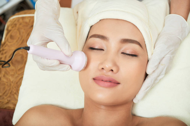 beautician hands making ultrasound facial procedure while pretty Asian client lying on treatment table with closed eyes - Foto, Bild