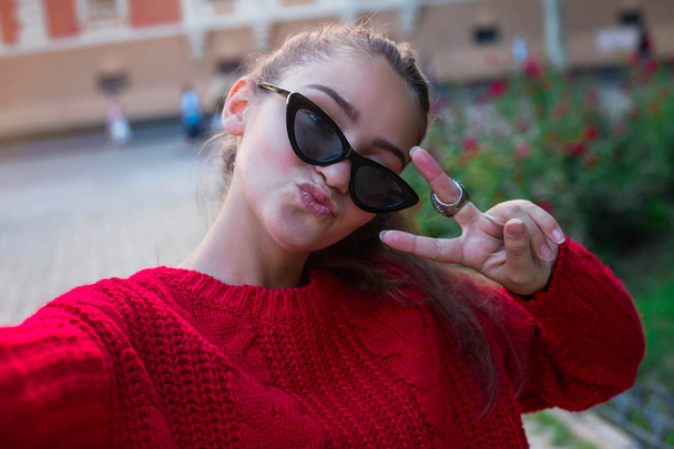 Young girl take selfie from hands with phone on spring city street. Urban life concept.Closeup selfie-portrait student of attractive girl in sunglasses with long hairstyle in city - Photo, Image