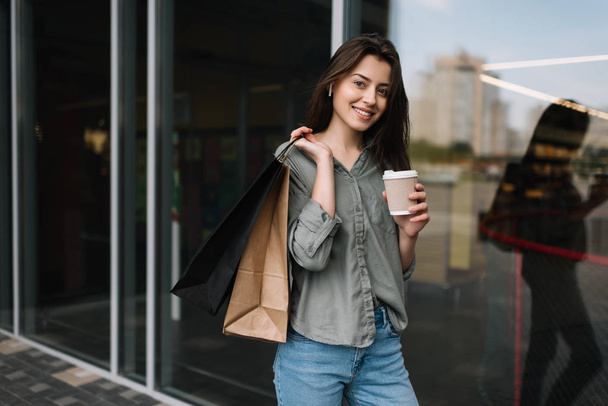 Милая женщина с красивой улыбкой идет по улице с сумками и чашкой кофе возле супермаркета или торгового центра. Молодая кавказская деловая женщина ждет такси на открытом воздухе
. - Фото, изображение