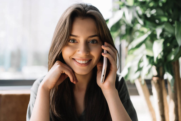Smiling attractive woman speaks on mobile phone in modern coffee shop. Young beautiful female using smartphone, calling taxi indoors - Photo, Image