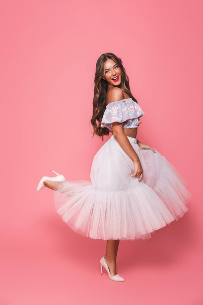 Full length portrait of positive excited woman 20s in beautiful skirt dancing and raising leg isolated over pink background in studio - Foto, Bild