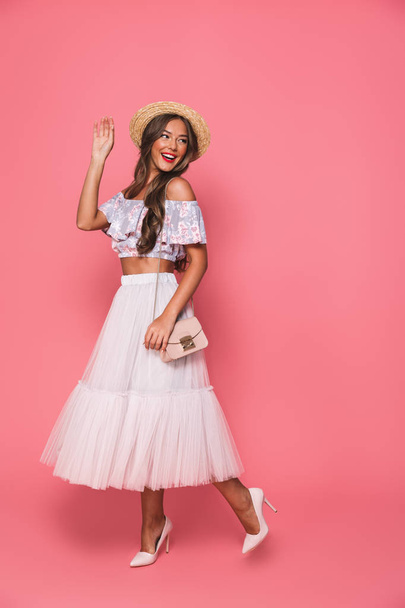 Full length image of brunette beautiful woman 20s wearing straw hat and fluffy skirt smiling and waving hand aside isolated over pink background in studio - Foto, Imagem