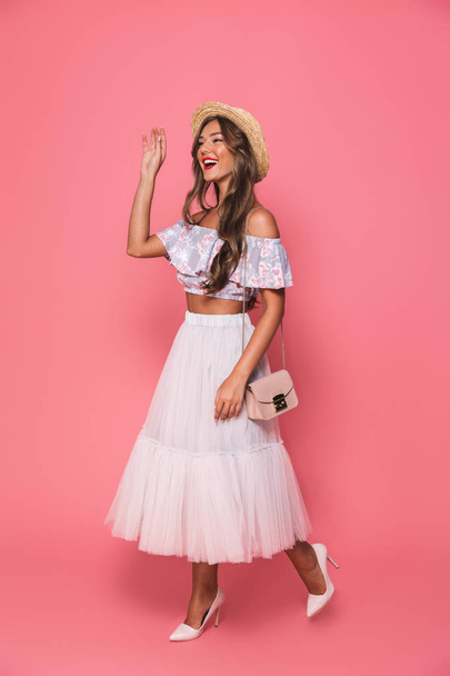 Full length portrait of attractive young woman 20s wearing straw hat and fluffy skirt smiling and waving hand aside isolated over pink background in studio - Photo, image
