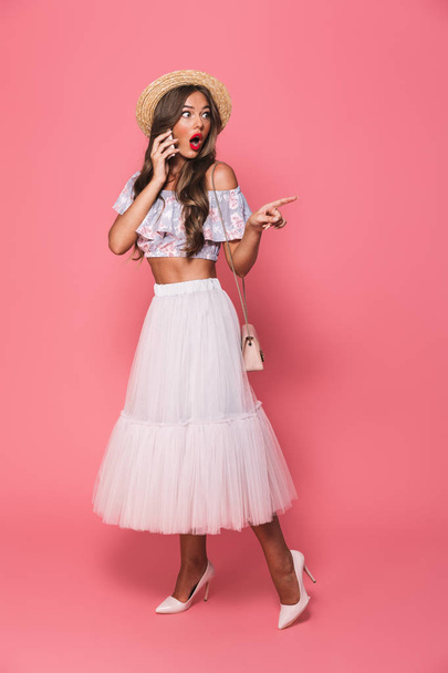 Full length portrait of amazed caucasian woman 20s wearing straw hat and fluffy skirt smiling and talking on cell phone isolated over pink background in studio - Photo, Image