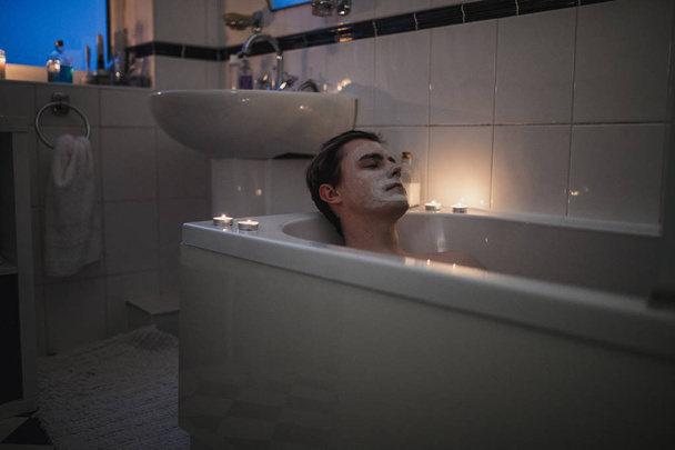 Молодий чоловік розслабляється у ванні з маскою для обличчя і свічками навколо нього
. - Фото, зображення