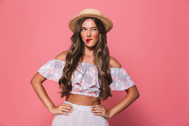 Portrait of seductive european woman 20s wearing straw hat biting lips and holding arms on waist isolated over pink background in studio - Foto, immagini