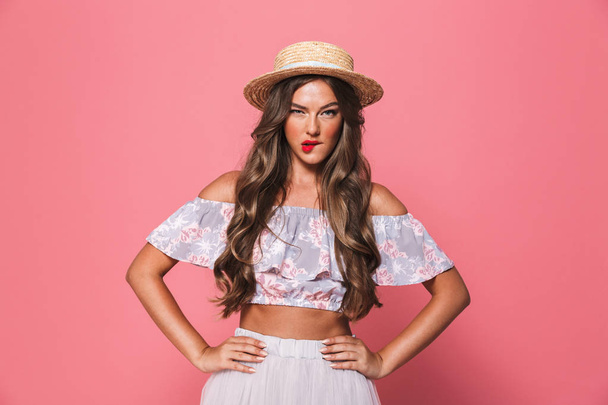 Portrait of serious pretty woman 20s wearing straw hat biting lips and holding arms on waist isolated over pink background in studio - Foto, afbeelding