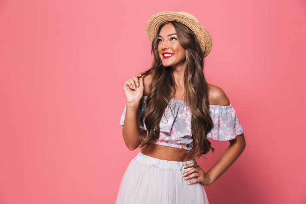 Portrait of positive glamour woman 20s wearing straw hat smiling and touching her beautiful brown hair isolated over pink background in studio - Photo, Image