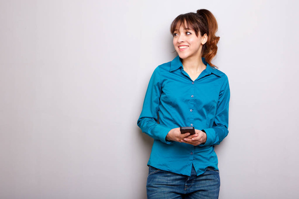 Porträt einer jungen Frau, die mit Handy vor grauem Hintergrund lächelt - Foto, Bild