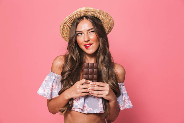 Portrait of a lovely young girl in summer clothes holding chocolate bar over pink background - Foto, immagini