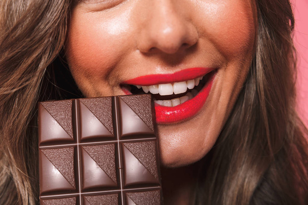 Close up portrait of a lovely young girl biting chocolate bar over pink background - Foto, Imagem