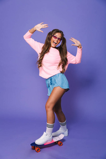 Full length portrait of a happy girl in sweatshirt posing on skateboard isolated over violet background - Zdjęcie, obraz