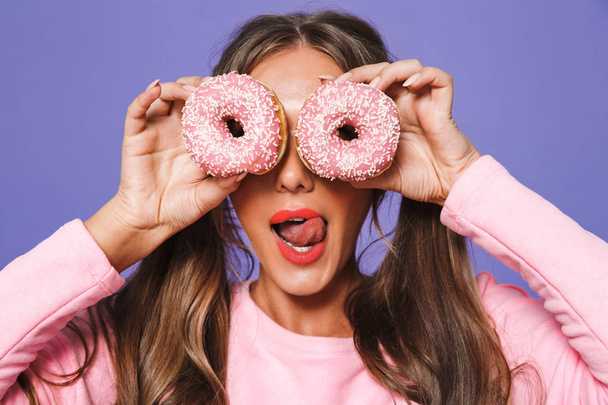 Close up portrait of a cheerful girl in sweatshirt posing with donuts at her face isolated over violet background - Foto, Bild