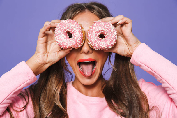 Close up portrait of a funny girl in sweatshirt posing with donuts at her face isolated over violet background - Foto, Imagem