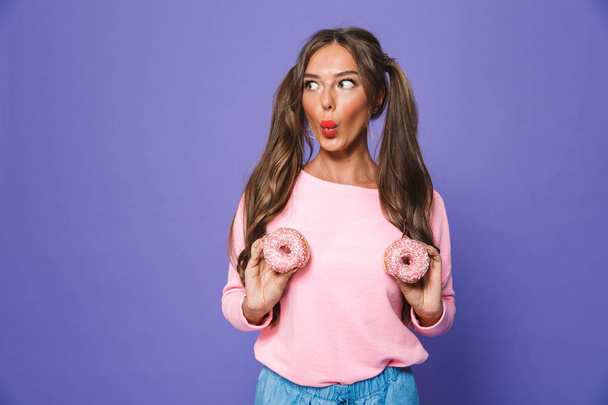 Portrait of a funny girl in sweatshirt posing with donuts isolated over violet background - Foto, imagen