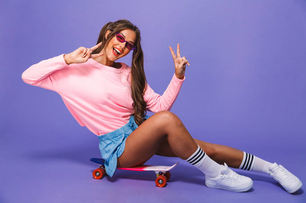 Portrait of a cheerful girl in sweatshirt posing on skateboard isolated over violet background, showing peace - Foto, immagini