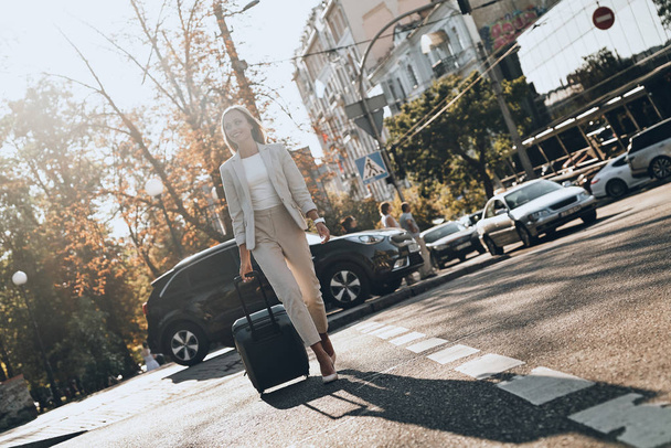 Confident business expert. Full length of young woman in suit pulling luggage and smiling while walking outdoors - Fotografie, Obrázek