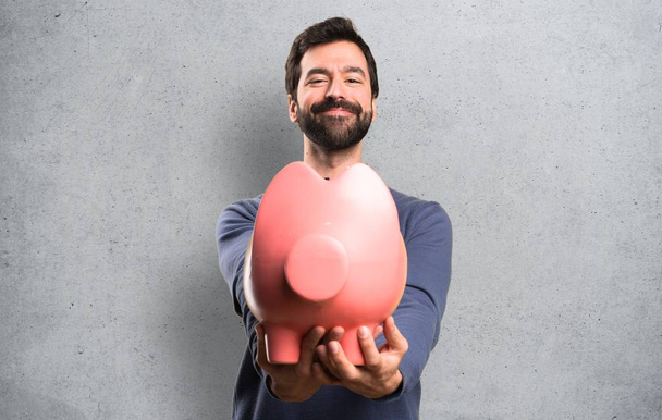 Feliz guapo morena hombre con barba sosteniendo un piggybank en texturizado fondo
 - Foto, Imagen