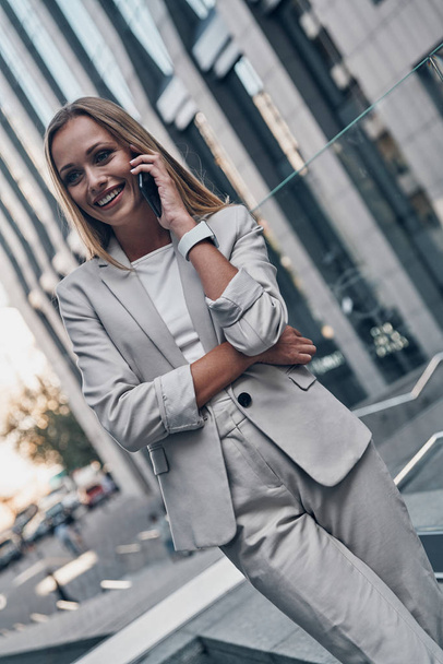 blonde beautiful woman in suit talking on phone and smiling while standing outdoors - Foto, afbeelding