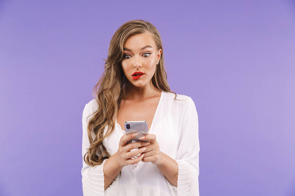 Photo of young brunette woman 20s with long curly hairstyle wearing dress holding and typing on smartphone isolated over violet background - Valokuva, kuva