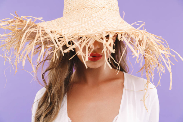 Photo of young fashion woman 20s wearing dress hiding her face with big straw hat and looking at camera isolated over violet background - Фото, зображення