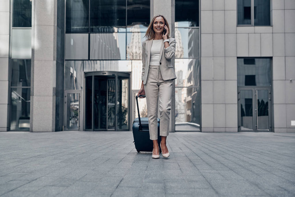 beautiful blonde business woman in suit pulling luggage while walking outdoors and talking on mobile phone  - Фото, изображение