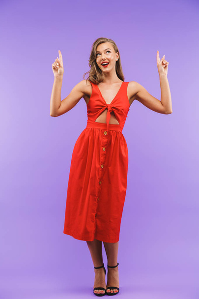 Full length portrait of a pretty young woman in red dress posing while pointing fingers up isolated - Фото, зображення