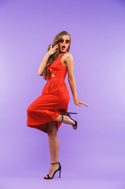 Full length portrait of fashion brunette woman 20s wearing red dress and sunglasses wondering or expressing surprise standing isolated over violet background - Фото, зображення