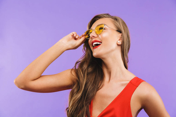 Image closeup of fashion brunette woman 20s wearing red dress and sunglasses smiling and looking aside at copyspace standing isolated over violet background - Foto, Imagem