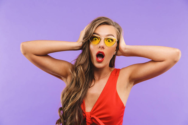 Photo of scared or shocked woman 20s wearing red dress and sunglasses screaming and covering ears with hands standing isolated over violet background - Foto, Imagem